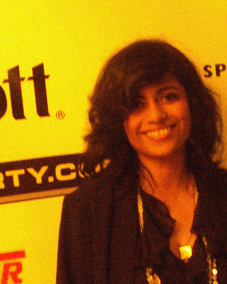 Night portrait, before: a smiling woman in a dark jacket against a yellow backdrop, with speckles in her hair